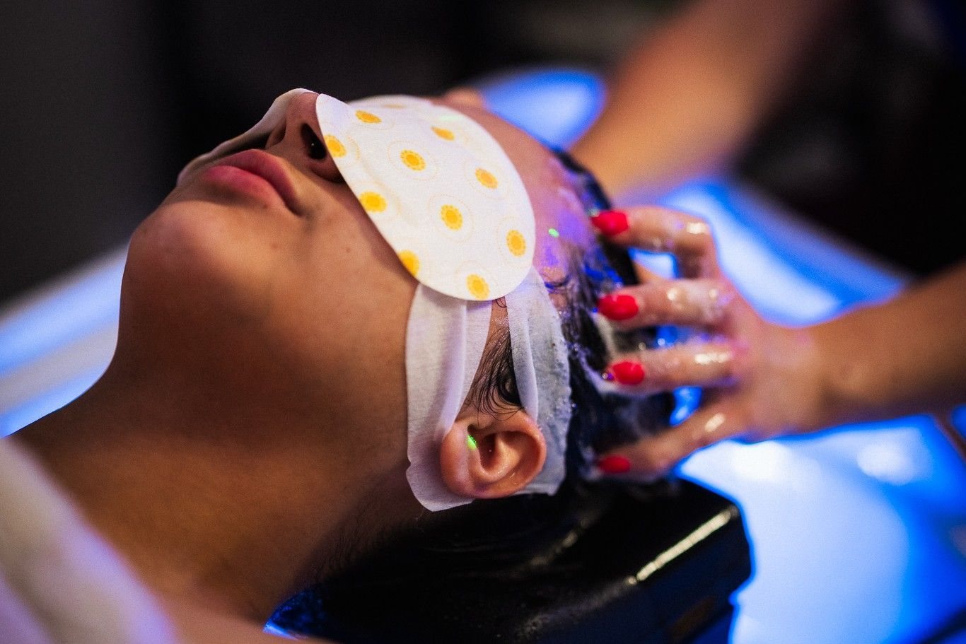 A client wearing a dotted eye mask while hands massage her scalp under blue light
