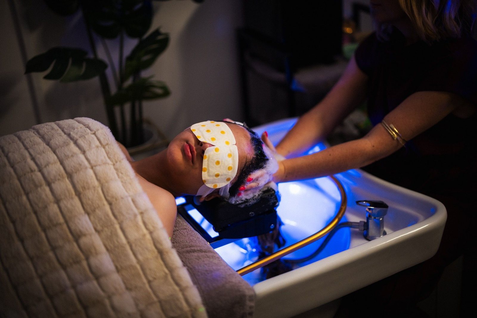 A client on the treatment bed with an eye mask, her head resting over the blue-lit basin